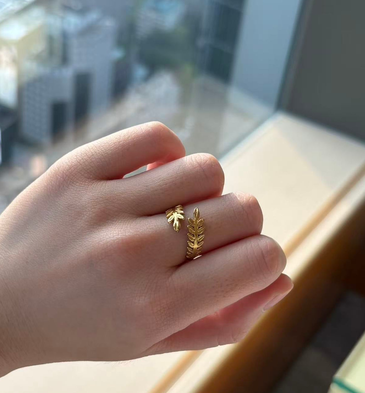 Hand wearing a gold ring with a leaf design in front of a window with a cityscape view.